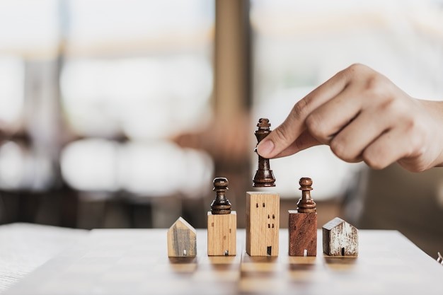 Mão de homem de negócios pegando em peças de xadrez posicionadas em cima de edifícios e casas de brinquedo, representando a dificuldade de encontrar estratégias para não perder imóvel com dívida condominial.