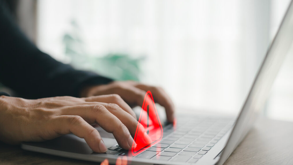 Pessoa utilizando um notebook com um triângulo vermelho com um sinal de exclamação, representando a importância de agir com cautela ao receber a notificação de leilão de imóvel.